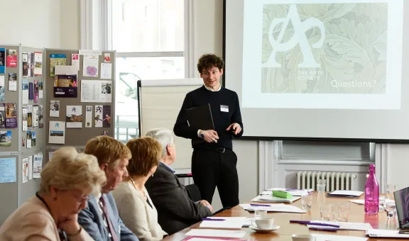 Staff member Joseph Little stands in front of a projector screen and leads a training session with 4 Arts Society Members listening